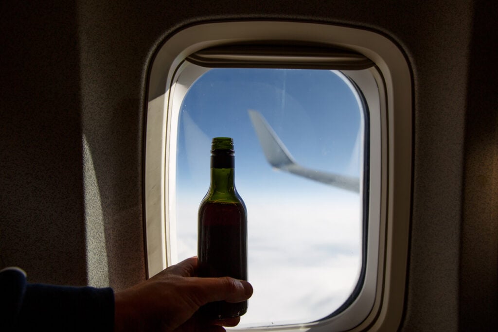 A hand holding a bottle against an airplane window with a view of the wing and clouds outside.