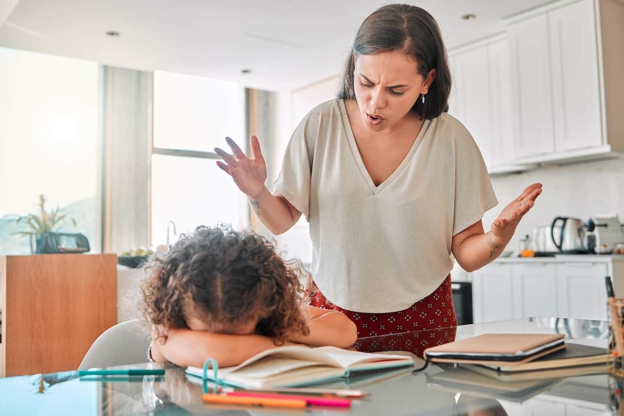 A frustrated woman gesturing while a child with their head down appears upset at a kitchen table with books and pencils.