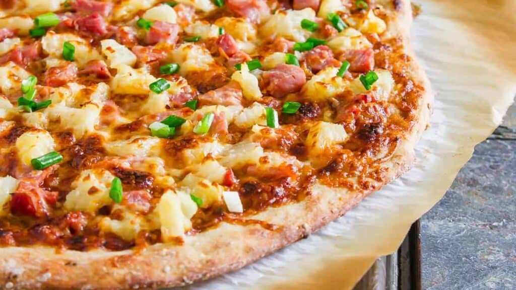 A close-up of a hawaiian pizza topped with ham, pineapple, cheese, and green onions.