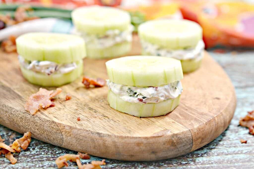 Sliced cucumbers with a creamy filling on a wooden cutting board, surrounded by crumbled bacon and vegetables.
