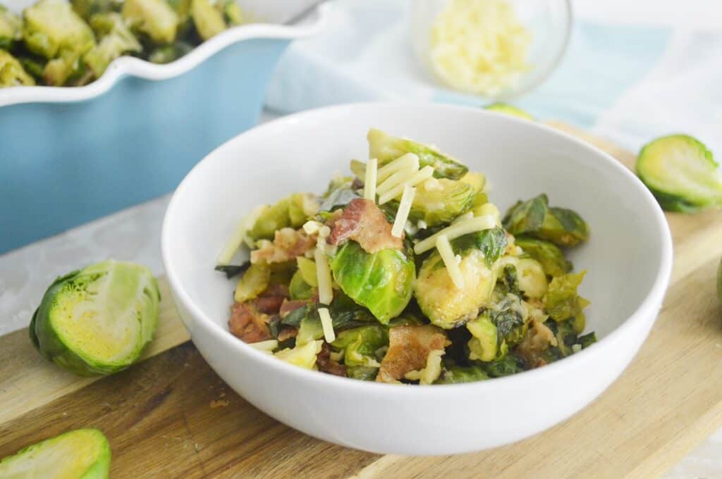 A bowl of brussels sprouts with bacon and shredded cheese on a wooden surface, with some raw brussels sprouts to the side.
