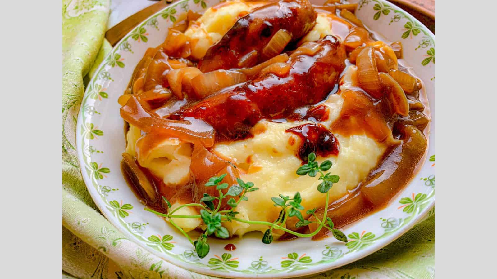 A plate of mashed potatoes topped with sausages and caramelized onions in a rich brown gravy. A sprig of fresh thyme garnishes the dish. The plate sits on a green patterned cloth.