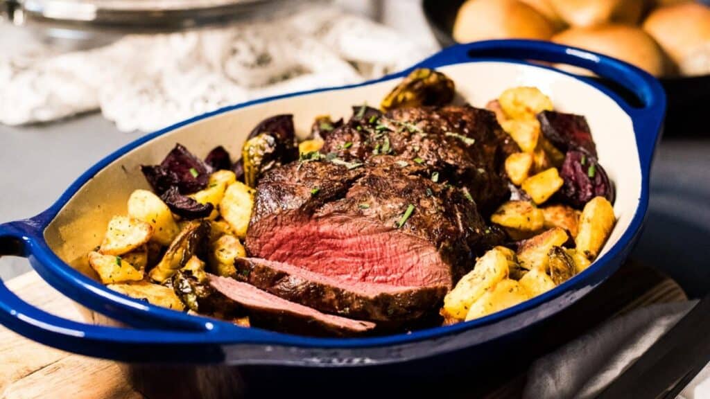 Roasted beef with herbs served with roasted potatoes and vegetables in a blue casserole dish.