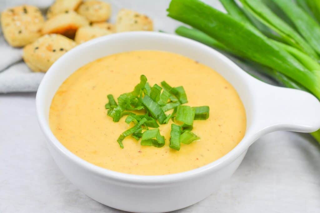 A bowl of creamy soup garnished with chopped green onions, accompanied by crackers on a kitchen countertop.