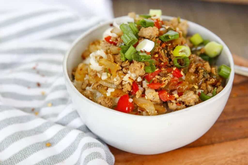 A bowl of stir-fried rice with vegetables, ground meat, and topped with chopped green onions and crushed nuts.