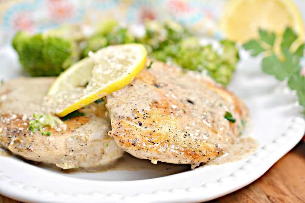 Grilled chicken breasts garnished with lemon slices and herbs, served with broccoli on a white plate.