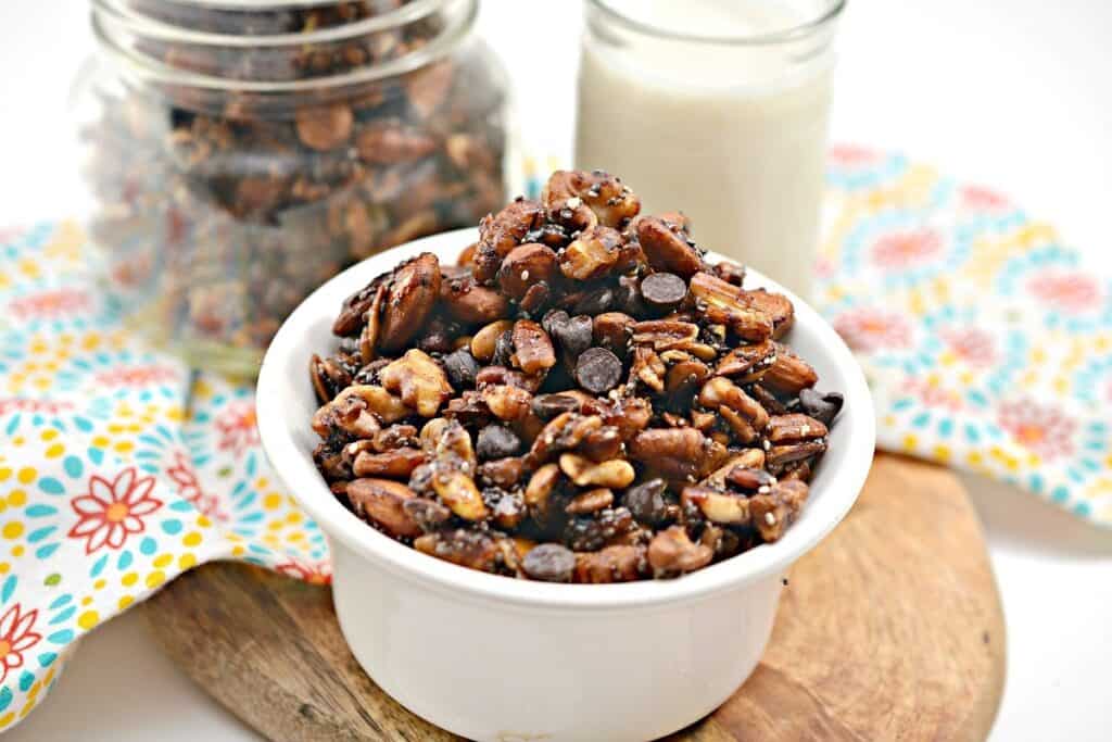 Bowl of mixed nuts and chocolate chips on a wooden surface with a glass of milk in the background.