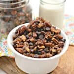 Bowl of mixed nuts and chocolate chips on a wooden surface with a glass of milk in the background.