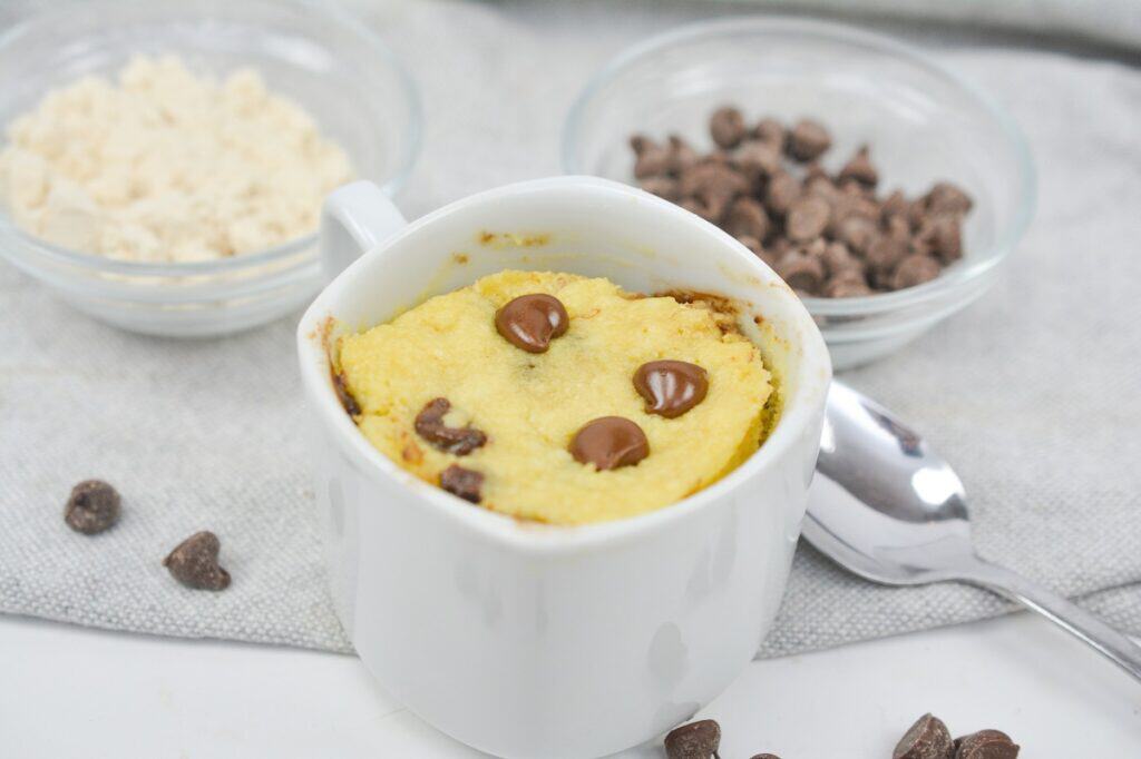 A freshly baked chocolate chip mug cake with ingredients in the background.