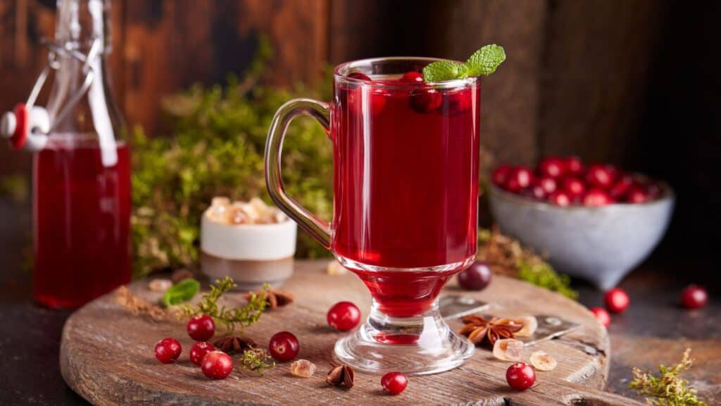 A glass of cranberry drink garnished with mint on a wooden board with cranberries and spices around.