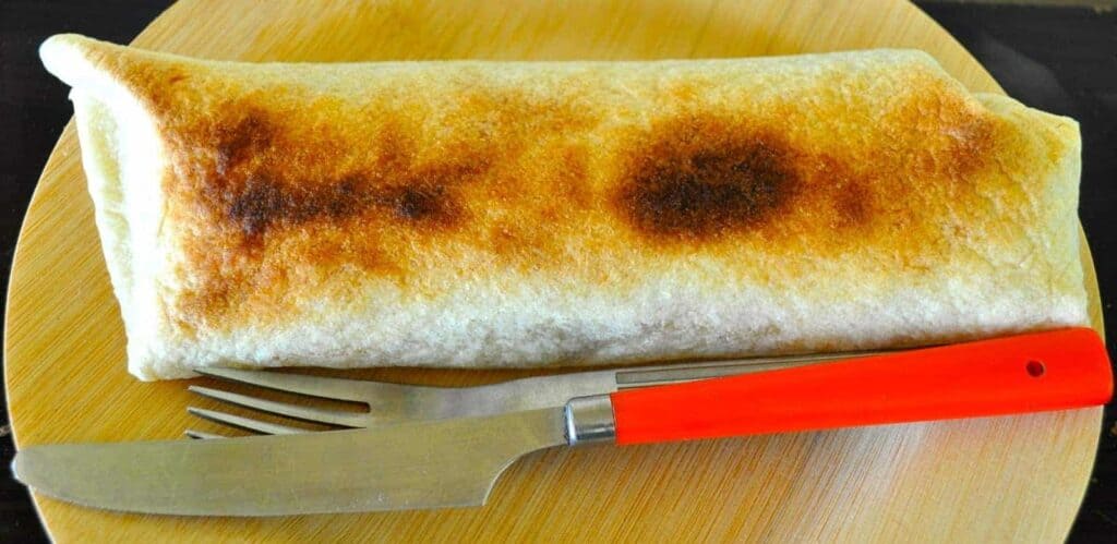 A toasted burrito on a wooden cutting board with a red-handled knife and fork beside it.
