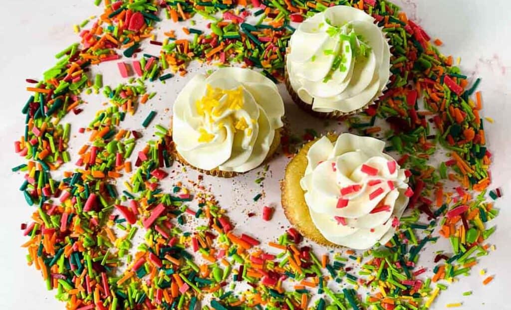 Cupcakes with white frosting and rainbow sprinkles on a white surface.