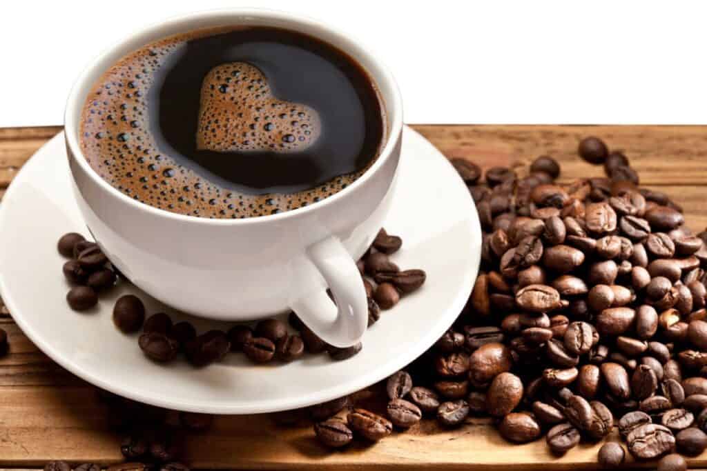 A cup of black coffee with a heart-shaped reflection on its surface, placed beside a pile of roasted coffee beans.