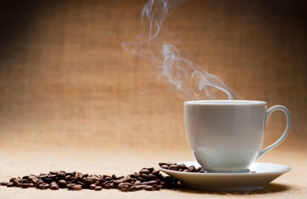 A white cup of hot coffee with steam rising, alongside coffee beans on a beige background.