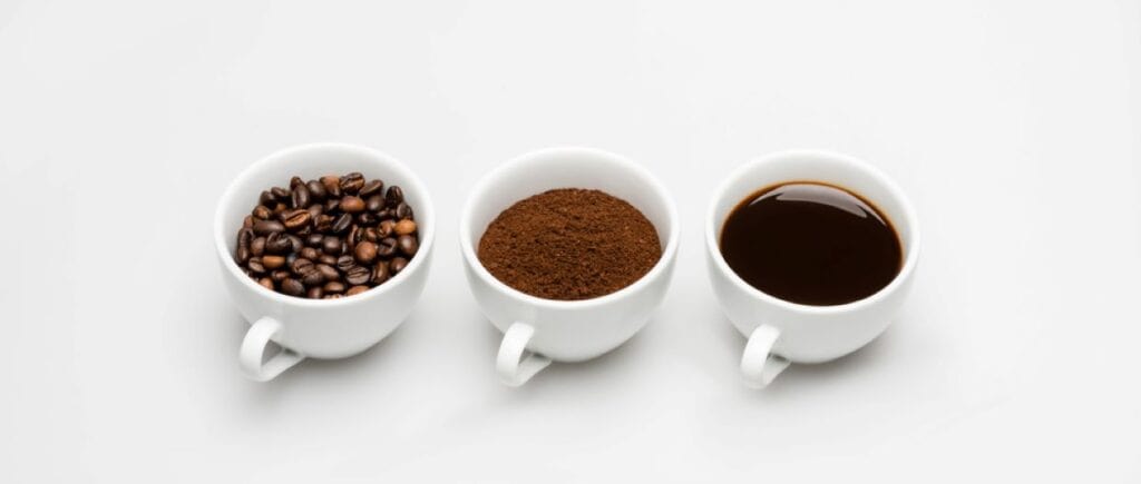Three white cups on a white surface, from left to right: one containing whole coffee beans, another with ground coffee, and the third filled with liquid coffee.