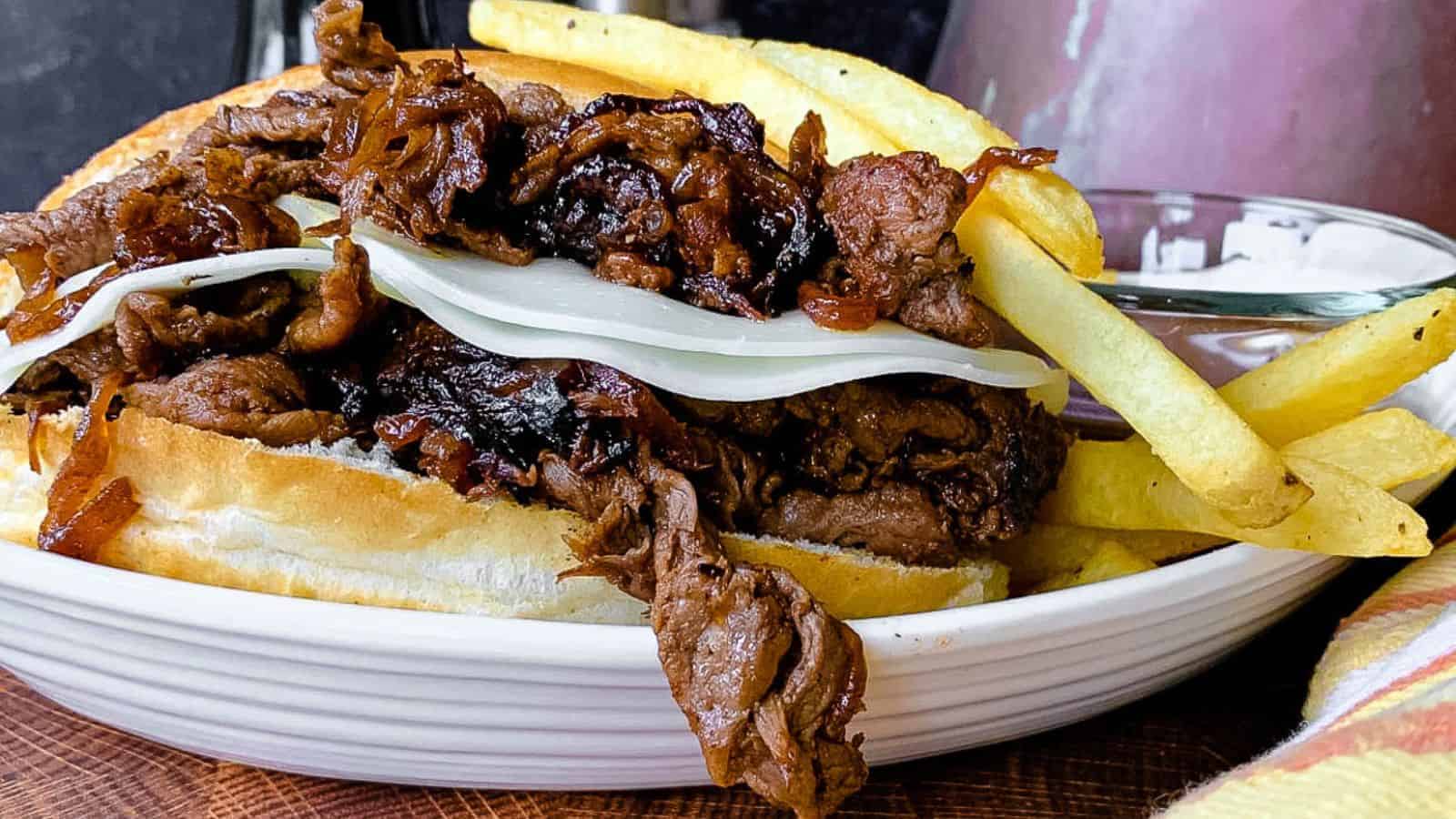 A shaved beef French dip sandwich on a platter with au jus and french fries.