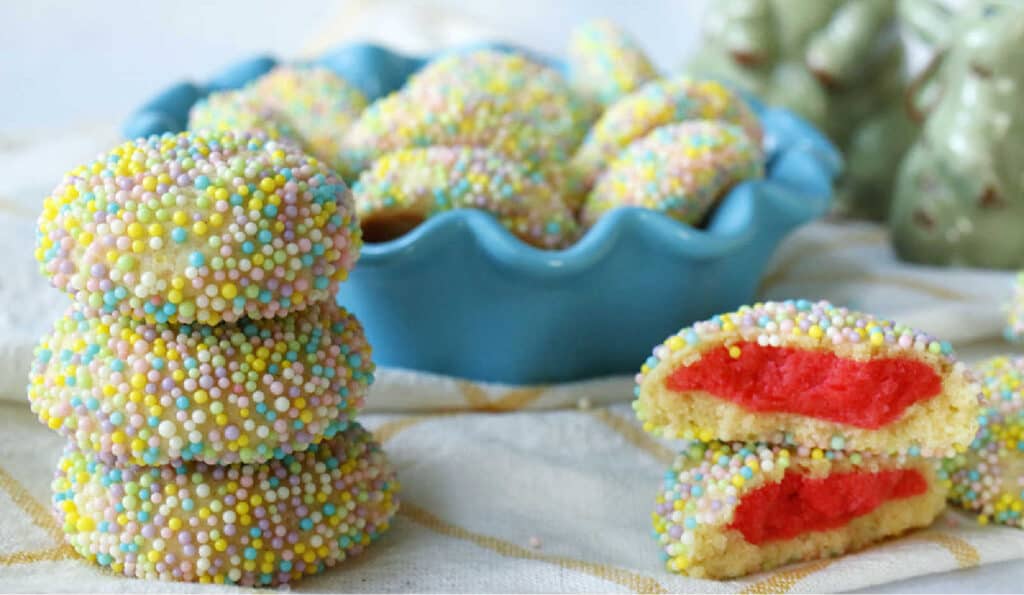 Colorful sprinkle-covered cookies in a blue bowl, one cut in half to reveal a red filling.
