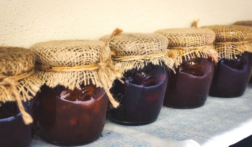 Jars of homemade preserves covered with burlap on a shelf.