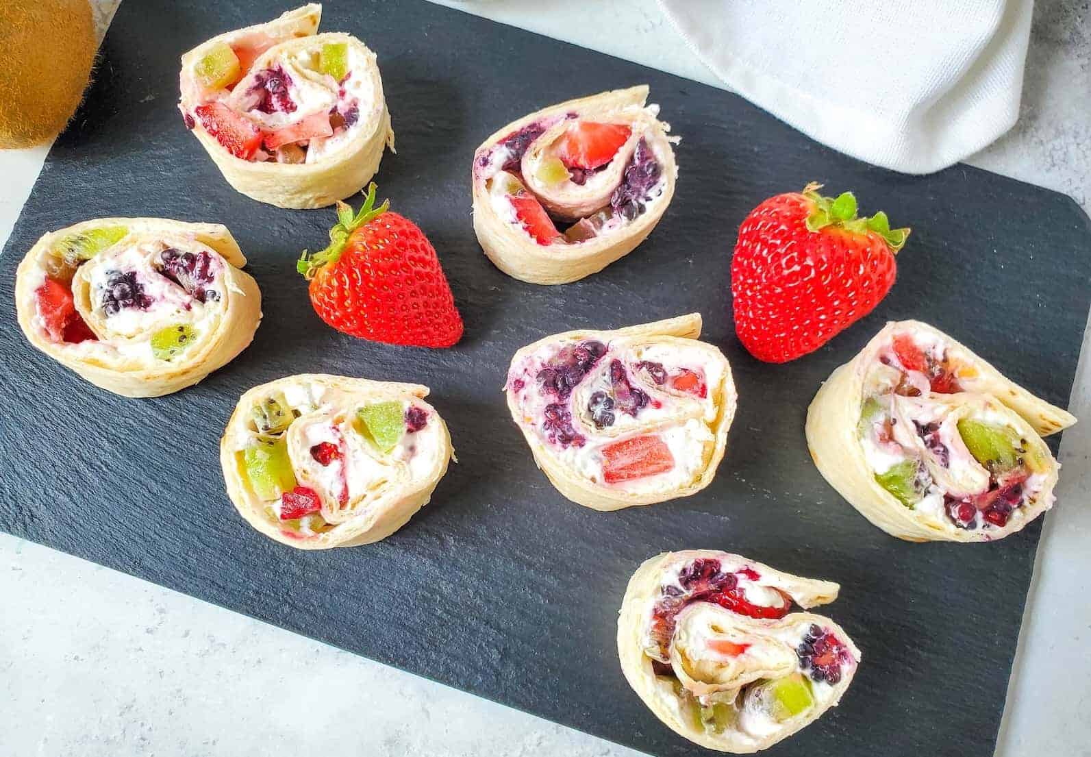 Sliced fruit and cream cheese pinwheel sandwiches arranged on a slate serving board with fresh strawberries.