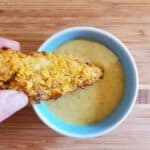 Image shows Hand dipping a corn flake crusted chicken tender into a bowl of sauce on a wooden table.