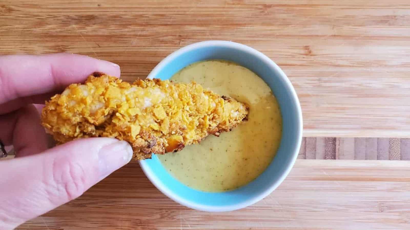 Image shows Hand dipping a corn flake crusted chicken tender into a bowl of sauce on a wooden table.