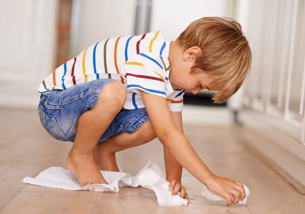 A young child crouches down to clean the floor with paper towels.