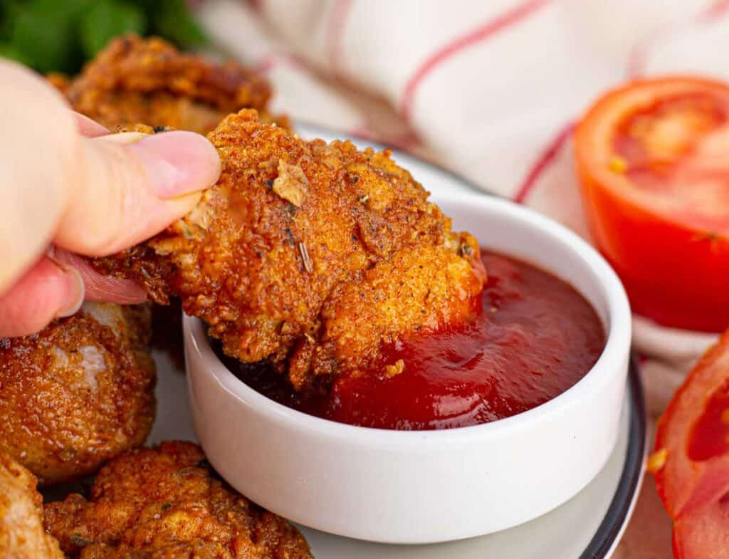 A hand dipping a piece of fried chicken into a bowl of ketchup.