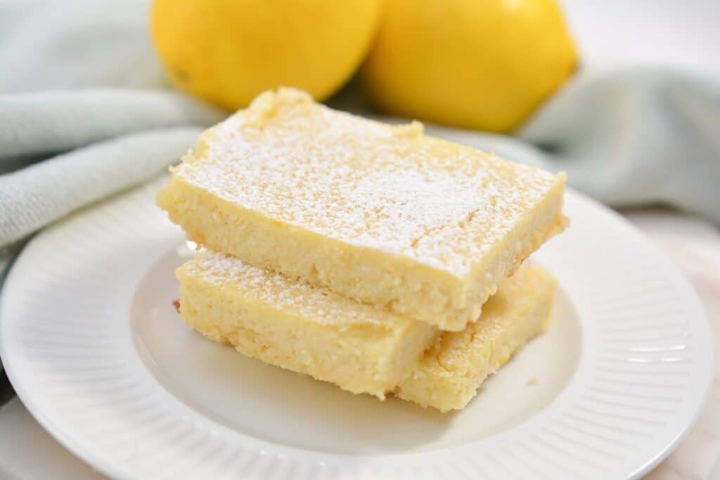 Three lemon bars dusted with powdered sugar on a white plate, with whole lemons in the background.