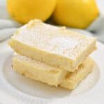 Three lemon bars dusted with powdered sugar on a white plate, with whole lemons in the background.