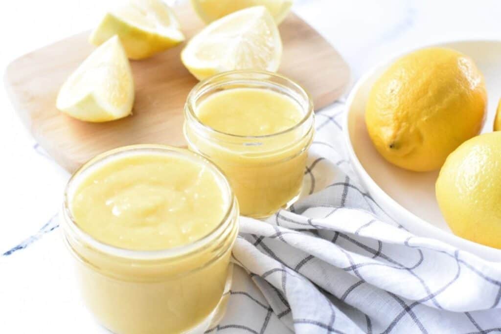 Freshly prepared lemon curd in glass jars with sliced lemons on a wooden cutting board in the background.