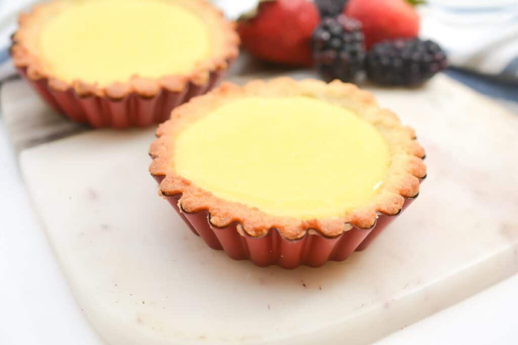 Two lemon tarts on a white surface with fresh berries in the background.
