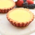 Two lemon tarts on a white surface with fresh berries in the background.