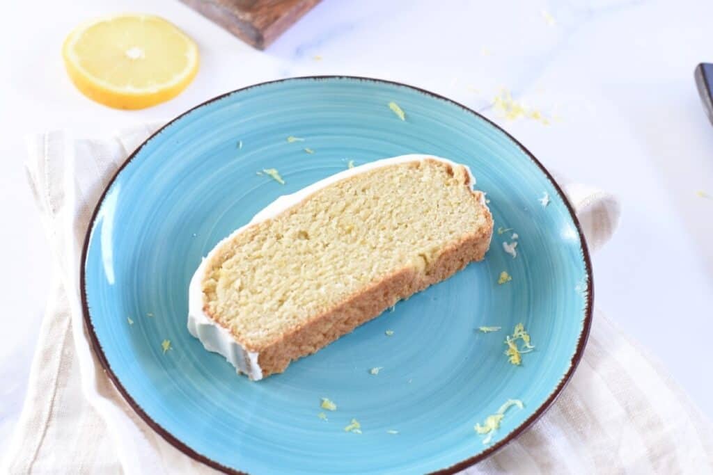 A slice of lemon cake with icing on a blue plate, garnished with lemon zest.