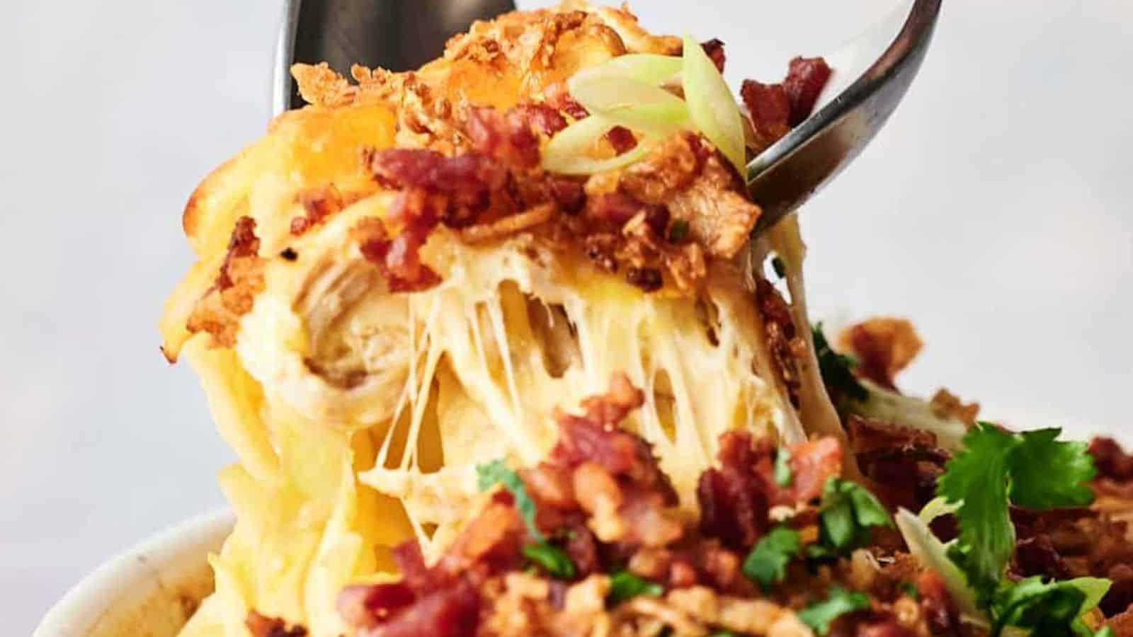A fork lifting a cheesy bite from a bowl of chicken casserole topped with bacon and breadcrumbs.