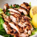 Grilled chicken breast slices served over a bed of greens with lemon wedges on the side.