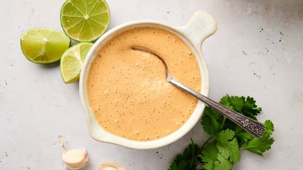 A bowl of creamy salad dressing with a silver spoon, accompanied by lime wedges and fresh herbs on a light surface.