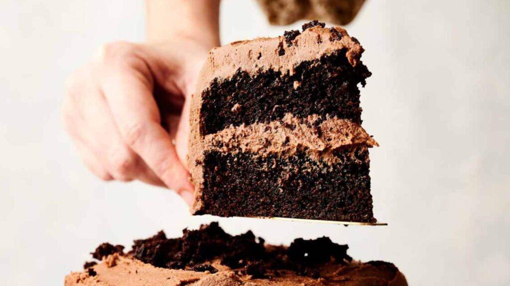 A hand holding a slice of chocolate layer cake with frosting.