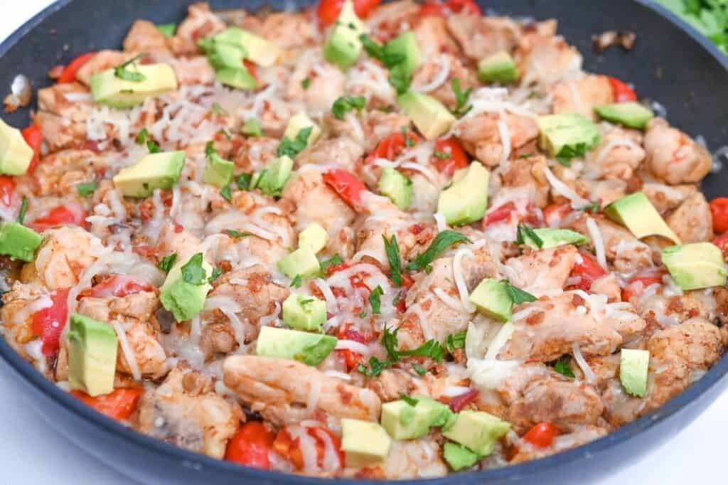 A skillet with cooked chicken, melted cheese, diced tomatoes, and chopped avocado, garnished with fresh herbs.