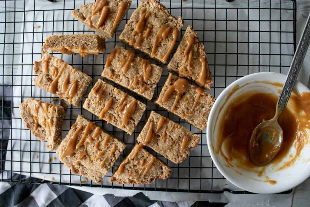 Slices of peanut butter bars on a wire rack drizzled with peanut butter, next to a bowl with extra peanut butter.