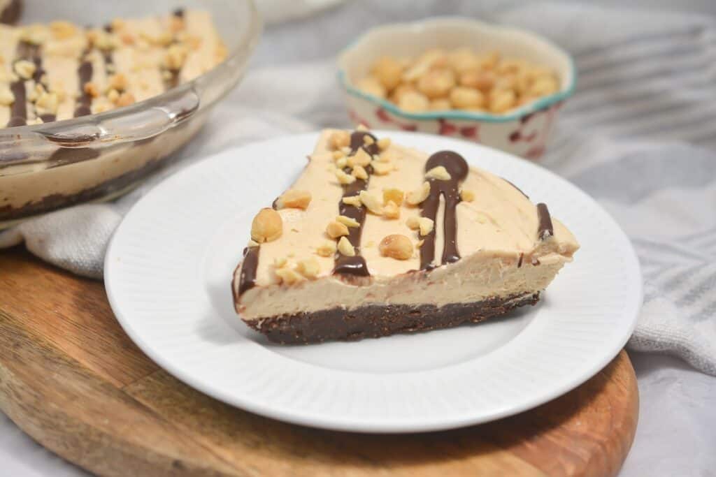 A slice of peanut butter pie with a chocolate drizzle and nut topping, served on a white plate with more of the dessert in the background.