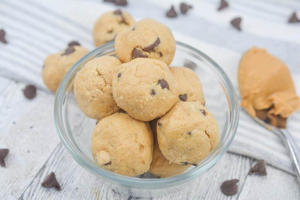 A bowl of peanut butter chocolate chip cookie balls on a wooden surface with chocolate chips and peanut butter in the background.