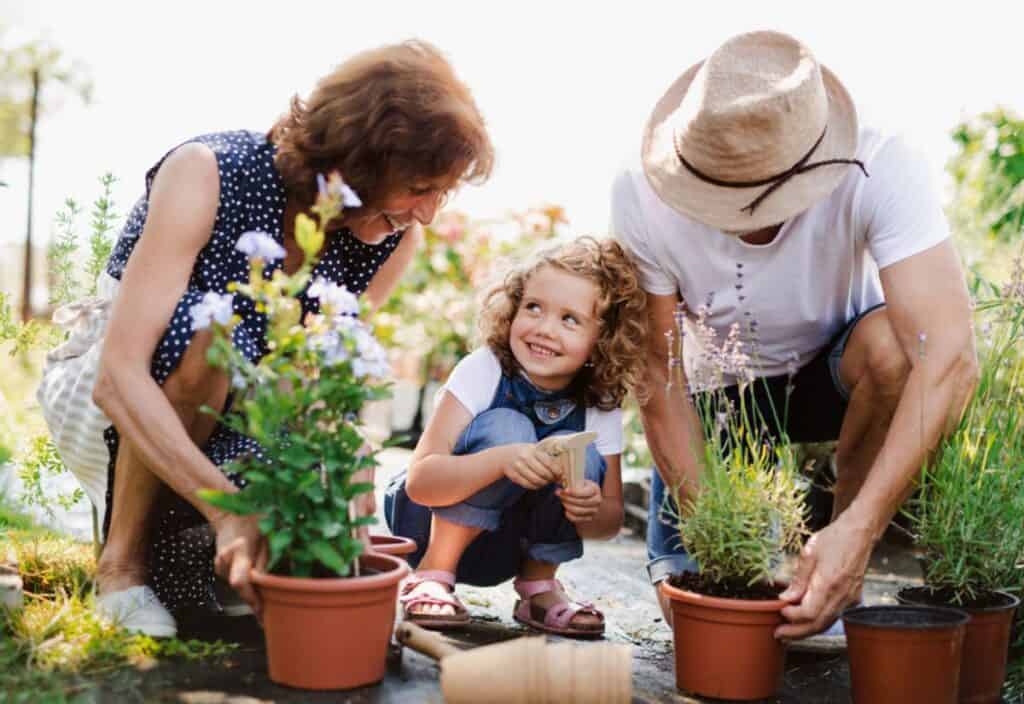 A young child assists two adults with gardening activities outdoors, watering plants in pots.
