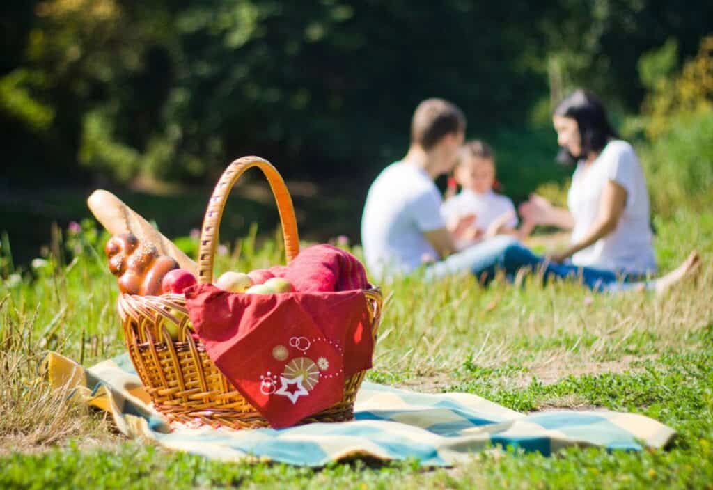 Picnic basket with bread and a red cloth on a blanket with a family in the background enjoying a picnic.