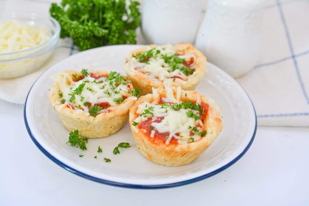 Three mini pizza cups on a white plate garnished with parsley, with shredded cheese and herbs in the background.