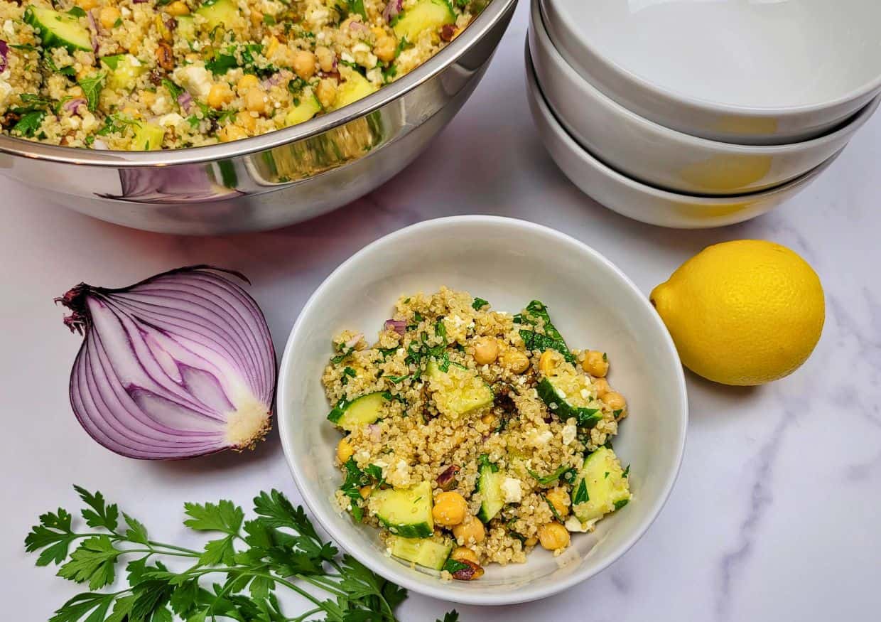 Quinoa salad with chickpeas, cucumber, and herbs served in a bowl, with a lemon, a red onion, and additional bowls on the side.