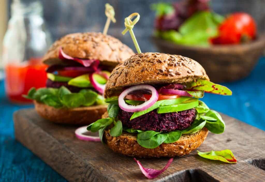 Two vegetarian burgers with beetroot patties and fresh vegetables on a wooden cutting board.