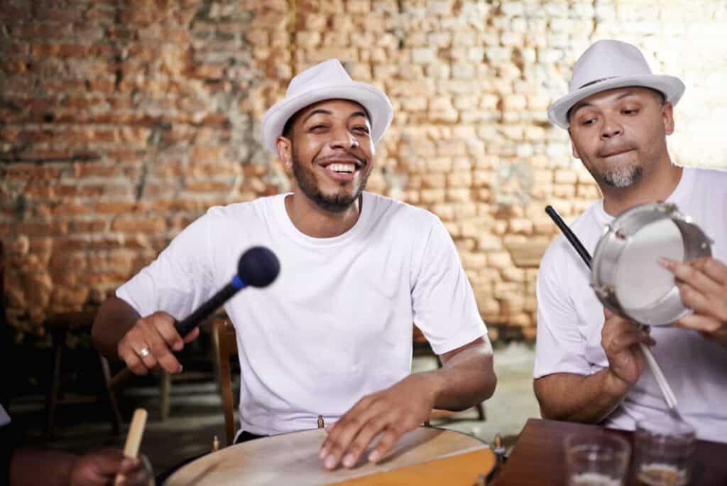 Two men in white hats and shirts playing percussion instruments and smiling.