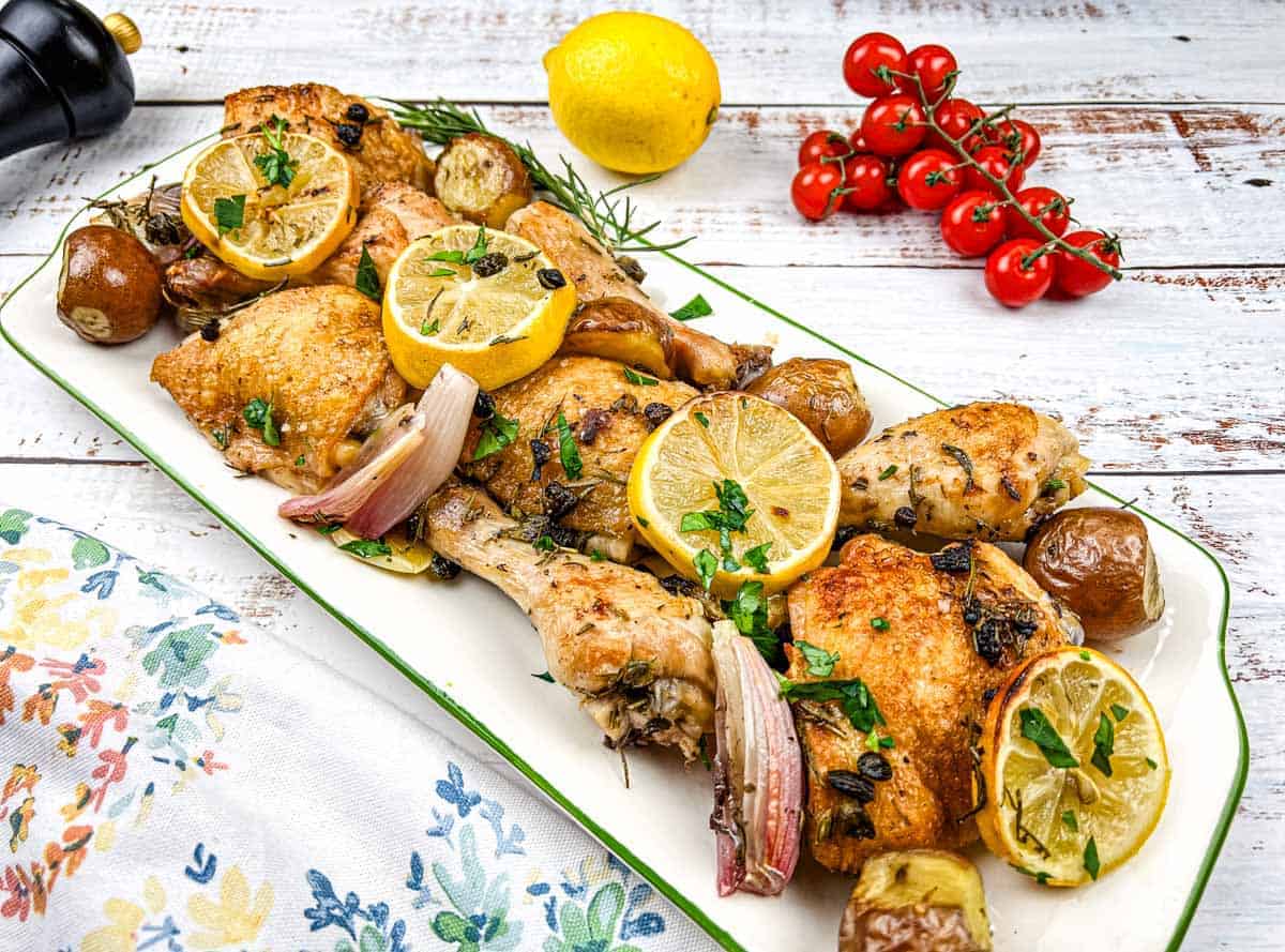 A platter of sheet pan lemon-rosemary chicken with lemon slices, herbs, and garnished with cherry tomatoes.
