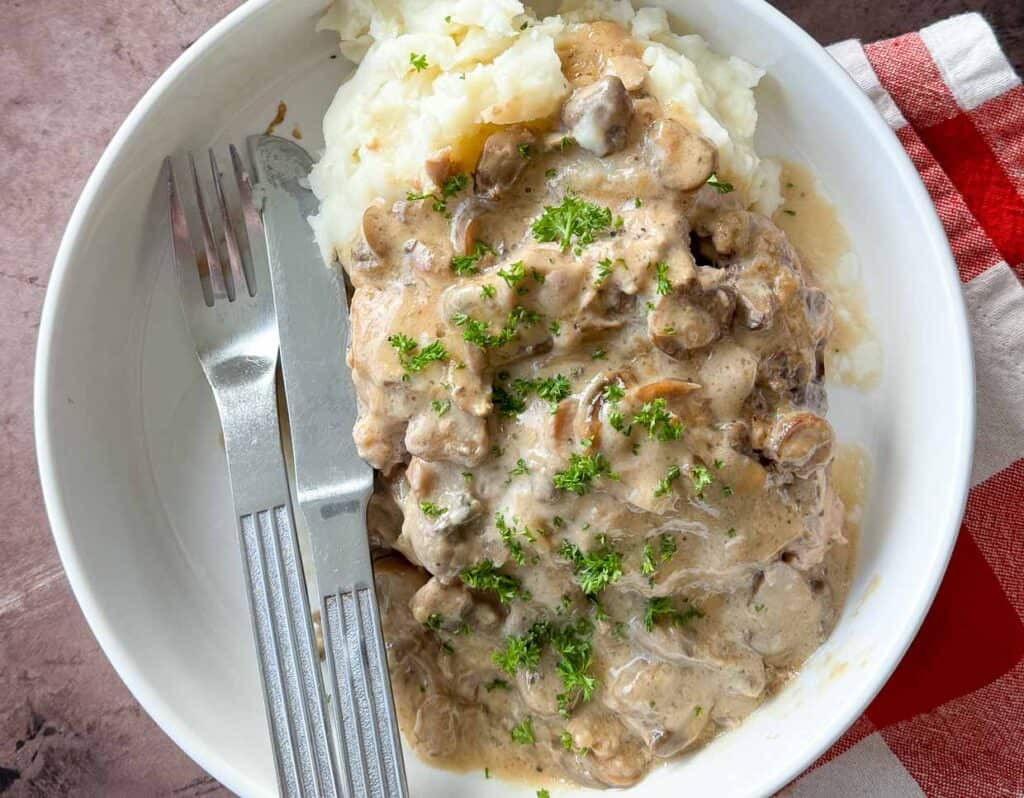 Plate of creamy mushroom beef stroganoff served over mashed potatoes, garnished with parsley, with a fork on the side.