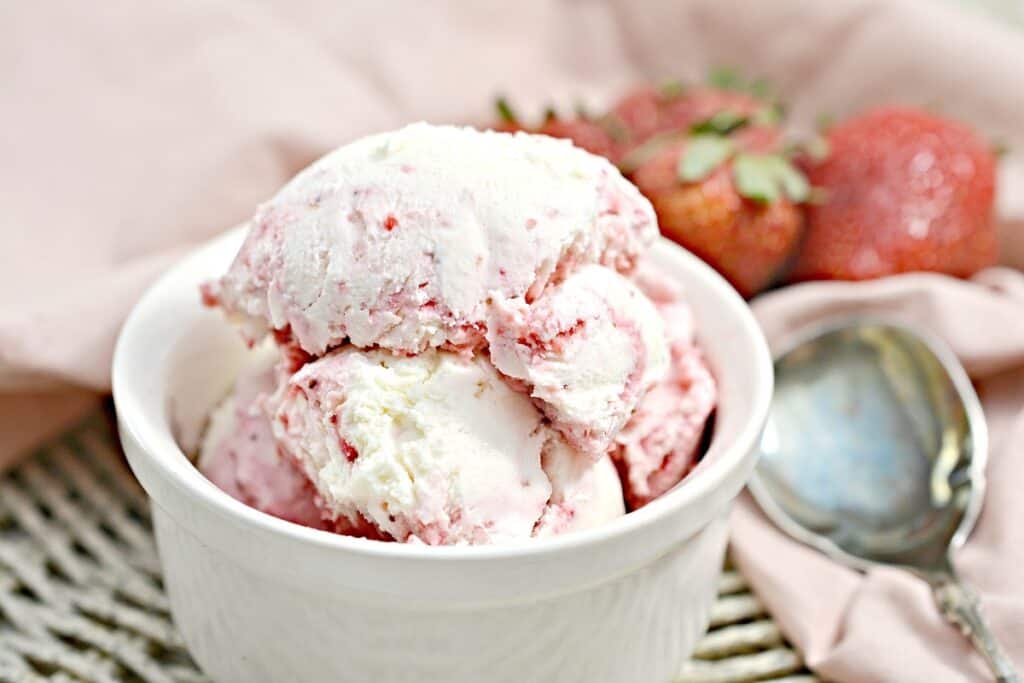 A bowl of strawberry ice cream with fresh strawberries on the side and a spoon resting on a napkin.